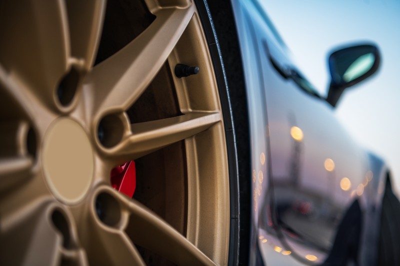 A close-up view of a gold alloy wheel with a red brake caliper, set against a blurred background of a car and soft lighting. The image highlights the design and details of the wheel.