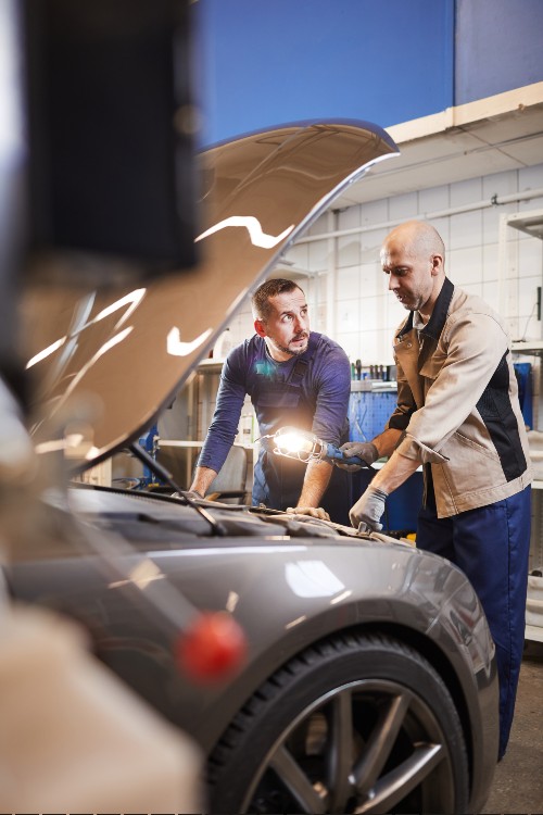 Two mechanics are working together under the hood of a car in a garage, examining the engine with a flashlight. The atmosphere is focused and collaborative.