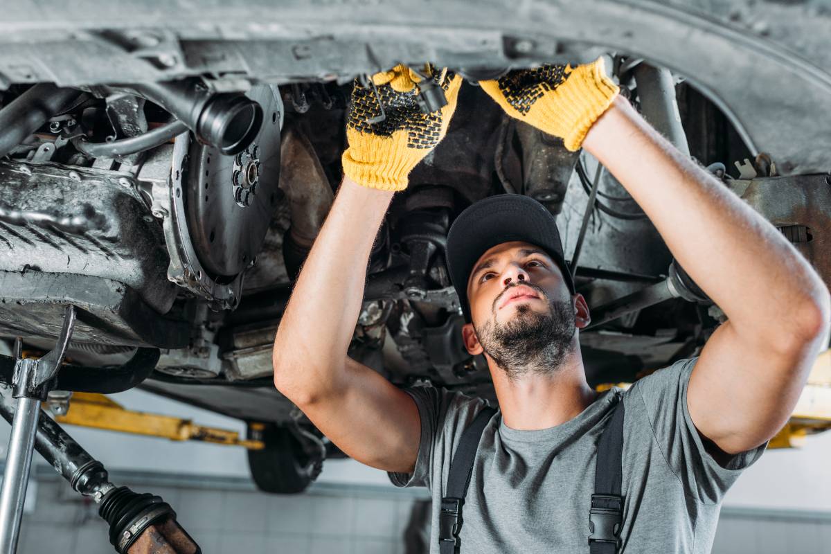 A mechanic works under a car, using tools to perform repairs while wearing gloves. The setting appears to be a garage or workshop.